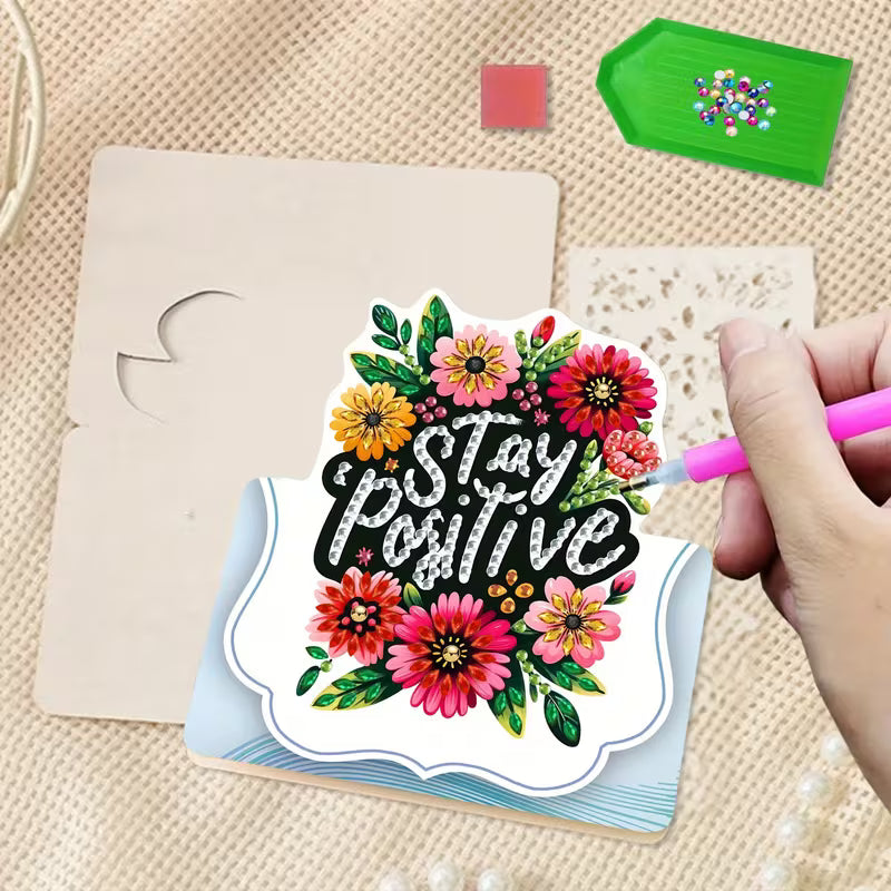 Hand holding a pink marker near a card with 'Stay Positive' and floral designs on a textured surface.