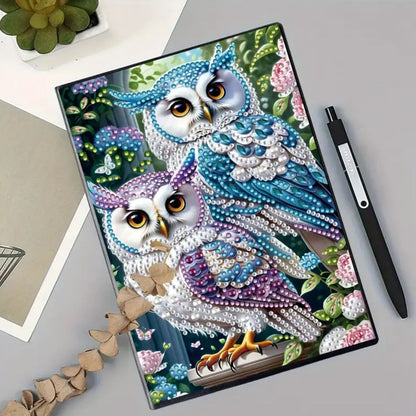 A decorated notebook with an owl design, featuring vibrant colors and rhinestones on the cover, placed on a table alongside a pen and a plant.