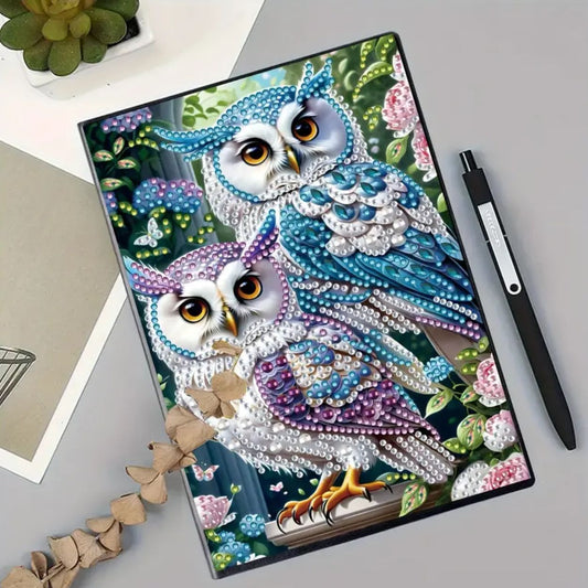 A decorated notebook with an owl design, featuring vibrant colors and rhinestones on the cover, placed on a table alongside a pen and a plant.