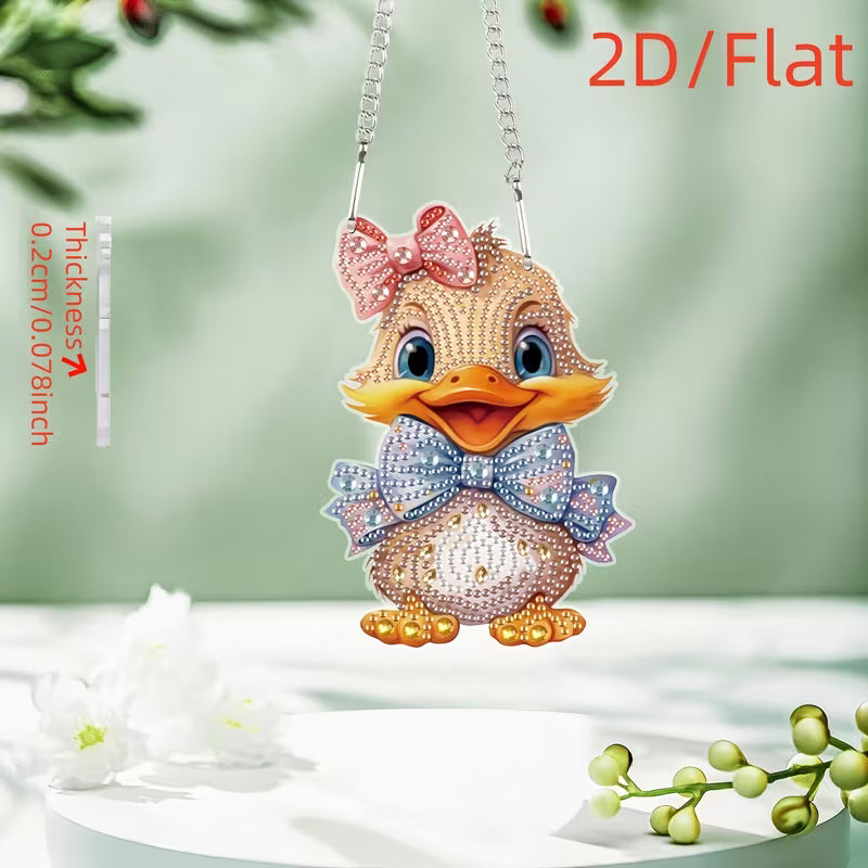 A 2D flat diamond painting pendant shaped like a cute duckling, decorated with pink, blue, and gold rhinestones, hanging from a silver chain against a soft green background with decorative white flowers.